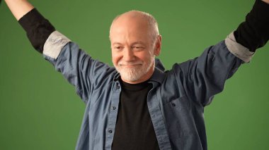 Üst düzey bir adam, katı yeşil bir altyapının önünde duruyor, açık elleri ve ifadesel yüzüyle çeşitli duygular sergiliyor, izleyiciyi doğrudan kendine çekiyor..