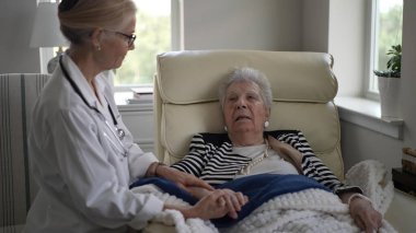 Olgun bir kadın evde bir sandalyede oturuyor ve bir sağlık uzmanı tarafından ziyaret ediliyor. El ele tutuşurken sıcak bir sohbete tutuştular..