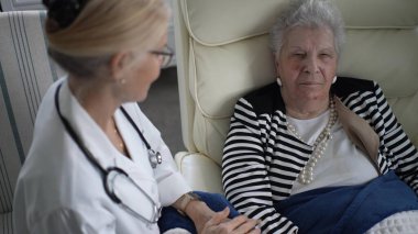 Olgun bir kadın, bir sağlık uzmanı sağlığını tartışırken rahat bir şekilde bir sandalyeye oturur. Konforlu ev atmosferi, kişisel bakımın önemini vurguluyor.
