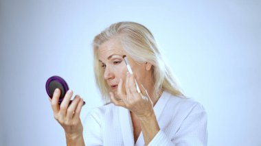 Beyaz bornozlu olgun bir kadın, spa 'nın rahatlatıcı atmosferinin tadını çıkarmak için fırçayla makyaj yapıyor. Neşeli ifadesi özenli anını yansıtıyor..