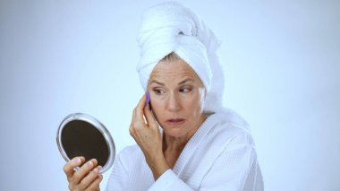 Spa elbisesi ve havlu giymiş olgun bir kadın ayna kullanarak makyaj yaparken gülümser, rahatlamayı ve kendine bakımı somutlaştırır, sakin, parlak bir ortamda..