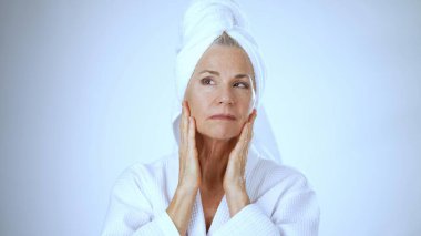 Olgun bir kadın beyaz bir bornoz ve havluya sarılmış bir spa ortamında neşeli bir şekilde gülümser. Huzurlu bir atmosferde rahatlama ve kendine bakımı kucaklar..