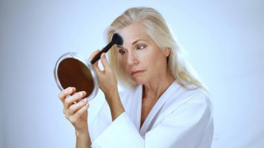 Beyaz cüppeli olgun bir kadın aynadaki yansımasını incelerken hafifçe gülümsüyor. Spa ortamı huzur ve rahatlama yayar..