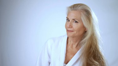 Spa bornozu giymiş olgun bir kadın uzun saçlarını tararken ışıl ışıl gülümsüyor. Huzurlu beyaz arka plan onun rahat ve neşeli atmosferine ekleniyor..