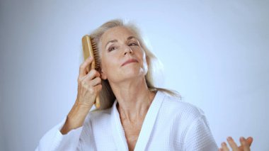 Beyaz cüppeli mutlu olgun bir kadın parmaklarını nazikçe uzun saçlarının arasında gezdirir. Huzurlu bir spa ortamında rahatlamanın tadını çıkarır..