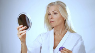 Beyaz cüppeli olgun bir kadın, sakin bir spa ortamında mor bir süngerle makyaj yaparken gülümsüyor. Yumuşak arka plan onun neşeli anını güçlendiriyor..