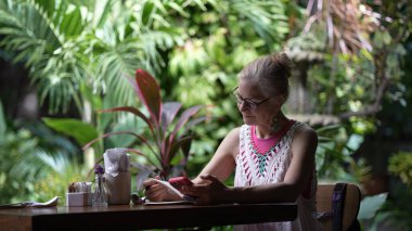 Olgun bir kadın kafeterya masasındaki işine odaklanır. O, verimli bir bahçe ortamında dikkatlice yazar, tatilde çalışan bir dijital göçebe olarak geçirdiği zamandan zevk alır..