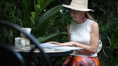 Olgun bir kadın bir kafe masasında oturur ve tropik bir ortamın tadını çıkarırken menüyü gözden geçirir. Tatildeki bir dijital göçebenin rahat yaşam tarzını somutlaştırıyor..