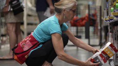 Winchester, VA, ABD - 06 28 2025: Olgun bir kadın evini yenileme projesini planlarken büyük bir hırdavatçıda boya ürünlerini inceliyor. Tadilatı için malzemeleri dikkatle seçer..