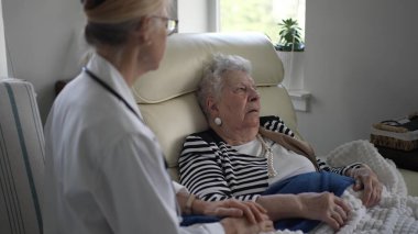 Yaşlı bir kadın evinde rahatça oturup, bir sağlık görevlisiyle sohbet ediyor. Sıcak bir atmosferde bir anlık bağ ve destek paylaşıyorlar..