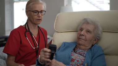 Bakıcı ve yaşlı bir kadın sıcak bir oturma odasında bir anı paylaşıyor. Kadın uzaktan kumandayla gülümserken sohbet ediyorlar..