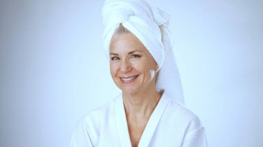 Olgun bir kadın rahatlatıcı spa gününün tadını çıkarırken mutlu bir şekilde gülümsüyor. Beyaz bir bornoz giyiyor ve kafasının etrafına parlak bir arka plan sarılı bir havlu var..