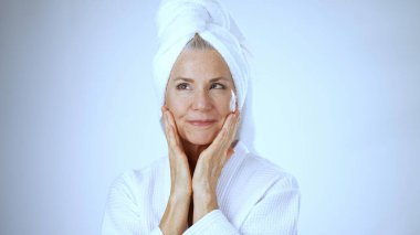 Kafasında havlu olan olgun bir kadın spa ortamında mutlu bir şekilde gülümsüyor. Temiz, beyaz arka plan onun gevşemiş ve neşeli tavrını arttırıyor..