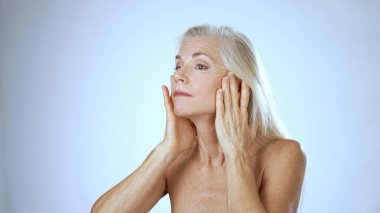 Sakinleştirici bir spa ortamında sarışın, olgun bir kadın yüzüne hafifçe dokunurken gülümsüyor. Yumuşak beyaz arka plan onun neşeli ifadesini arttırır..