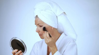 Beyaz cüppeli olgun bir kadın spa 'da aynanın önünde makyaj yaparken gülümsüyor. Huzurlu arka plan onun mutlu ve rahat tavırlarına ekleniyor..