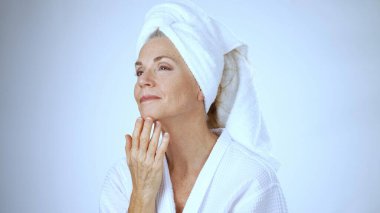 Olgun bir kadın kaplıcada dinlenirken mutlu bir şekilde gülümser, beyaz bir bornoz giyer ve temiz beyaz bir arka planda saçına havlu takar. Neşe ve sükunet saçıyor..