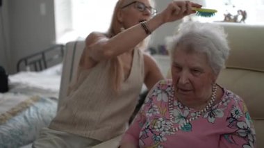 Olgun bir kadın, sıcak bir oturma odasında yaşlı annesinin saçlarını nazikçe tarar, samimi bir ziyaret sırasında bağlarının altını çizer..