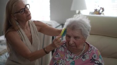 Olgun bir kadın, sıcak bir öğleden sonra ziyareti sırasında özen ve şefkatle dolu bir oturma odasında otururken yaşlı annesinin saçlarını nazikçe tarar..