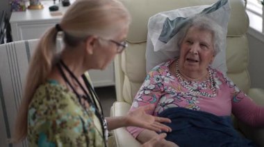 Yaşlı bir kadın, huzur içinde koltuğunda oturur ve bir hemşire huzur evinde rutin bir ziyaret sırasında arkadaşça sohbet ederken hayati organlarını kontrol eder..