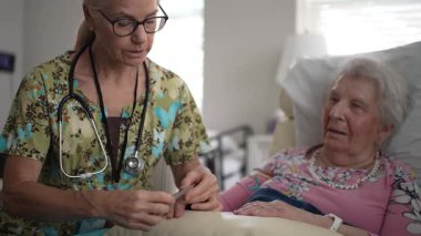 Bir hemşire yatakta oturan yaşlı bir kadının tırnaklarını dikkatlice törpülüyor. Paylaştıkları şefkatli bir anda atmosfer sakin..