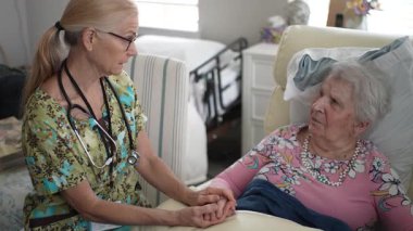 Bir hemşire yaşlı bir kadının elini tutarak huzur verici bir ortam yaratmak için huzur verici bir huzurevinde dostça bir konuşma yapar..
