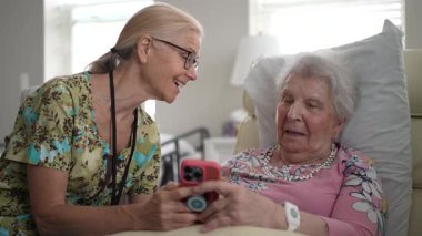 Hemşire ve yaşlı bir kadının portresi sıcak bir şekilde etkileşime geçiyor, yaşlılar yurduna yapılan bir ziyarette telefon kullanarak anılarını ve bilgilerini paylaşıyor..