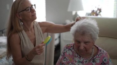 Olgun bir kadın, sıcak bir oturma odasında sıcak bir ziyaret sırasında sevgi ve ilgi göstererek yaşlı annesinin saçlarını nazikçe tarar..