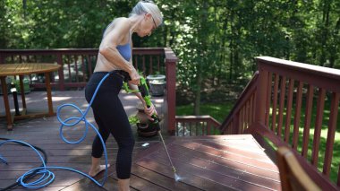Olgun bir kadın ahşap güvertesini temizlemeye çalışıyor. Basınçlı çamaşır makinesiyle. Arka bahçesinin güneşli bir günde taze ve davetkar görünmesini sağlıyor..