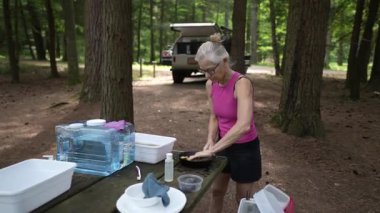 Olgun bir kadın, ağaçlarla çevrili bir kamp alanında bulaşık yıkıyor. Yakınlarda bir karavanı var. Açık havada huzurlu bir yaşam tarzı sergiliyor..