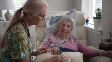 Bir hemşire yaşlı bir kadının oksijen seviyesini oksitölçer kullanarak dikkatle izler. Parlak bir odadalar, yaşlılar için sağlık hizmetleri ve desteklerini ön plana çıkarıyorlar..