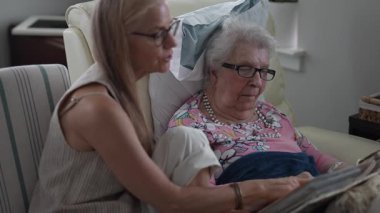 Huzurevinde, bir kız ve yaşlı annesi bir fotoğraf albümündeki mutlu anılarını paylaşırken gülümsüyor, birbirlerine eşlik ediyorlar ve gülüyorlar..