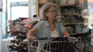 Yaşlı bir kadın markette geziniyor, içi yiyecek dolu bir alışveriş arabasını itiyor. Alışveriş tercihlerine ve çevresine odaklanmış görünüyor..