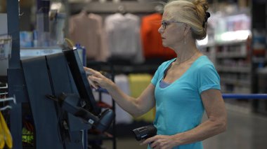 Olgun bir kadın, hırdavatçıda kendini keserken konsantre oluyor. Eşyalar satın alıyor ve elinde bir tarayıcı tutuyor..