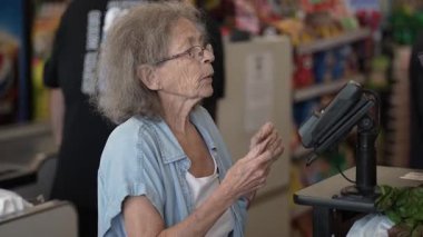 Gri saçlı yaşlı bir kadın kasanın önünde duruyor. Renkli ürünler ve eşyalarla dolu bir dükkanda alışverişini yapmaya hazırlanıyor..