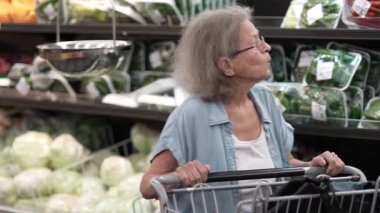 Yaşlı bir kadın bir alışveriş arabasıyla markette dikkatlice geziniyor, renkli ürünler arasında taze sebzeler arıyor..