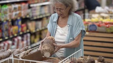 Üst düzey bir kadın alışveriş gezisi boyunca renkli meyve ve sebzelerle çevrili bir markette kök sebzeyi dikkatle inceliyor..