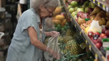 Yaşlı bir kadın marketten taze meyve ve sebze seçiyor. Seçtiği eşyaları alışveriş çantasına koyarken seçeneklerini dikkatlice inceler..