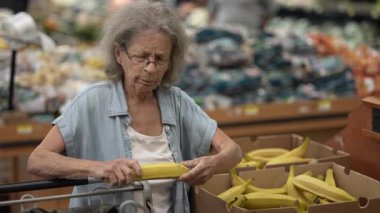 Gri saçlı yaşlı bir kadın alışveriş arabasını iterken kalabalık bir markette taze yiyecek seçiyor. Listesine ve yakındaki ürünlere odaklanıyor..