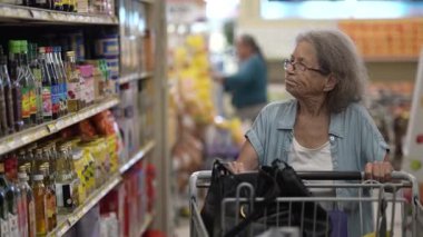 Yaşlı bir kadın, marketteki ürün raflarına dikkatlice bakarken alışveriş yaparken yemek seçeneklerini düşünen bir alışveriş arabasını itiyor..