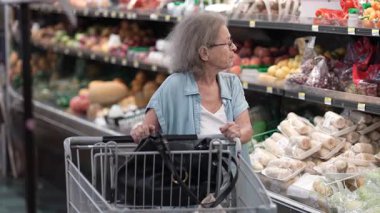 Yaşlı bir kadın bir marketin koridorlarında geziniyor, canlı vitrinlerde alışveriş arabasını iterken dikkatle taze ürünleri seçiyor..
