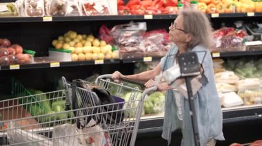 Kıdemli kadın markette taze meyve ve sebzelere dikkatlice bakıyor, alışveriş arabasını iterken yemekleri için seçimler yapıyor..