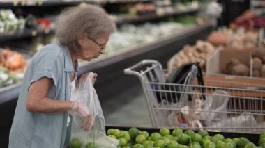 Üst düzey bir kadın alışveriş yaparken limonları dikkatlice inceler. Seçilen meyveyi çantasına doldurulmuş taze ürünlerle dolu bir mağaza koridoruna koyar..
