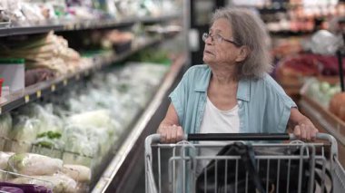 Yaşlı bir kadın alışveriş arabasını bir markete doğru itiyor ve raflardaki çeşitli sebzeleri inceliyor..