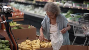Yaşlı bir yaşlı kadın taze meyve ve sebzelerle dolu canlı bir mağazadan muz alırken dikkatli bir şekilde muz seçiyor ve mevcut çeşitliliğin tadını çıkarıyor..