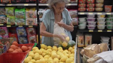 Yaşlı bir kadın, marketteki ürünler arasından taze limonları dikkatlice seçer. Alışverişe odaklanmış ve meşgul görünüyor..