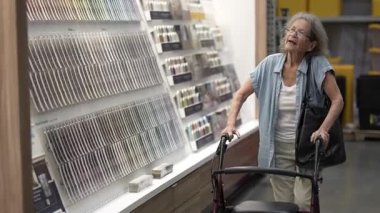 Yürüteç kullanan yaşlı bir yaşlı kadın, bir hırdavatçıda çok sayıda boya rengi seçerek ev dekorasyonu projesi için seçenekleri değerlendiriyor..