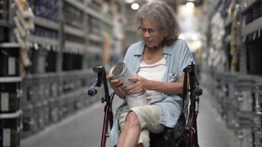 Kıdemli bir kadın bir hırdavatçıda renk seçeneklerine odaklanmış bir boya kutusunu inceliyor. Yürüyen bir tekerlekli sandalyede oturuyor. Etrafı çeşitli boya seçenekleriyle çevrili..