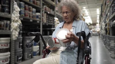 Yaşlı yaşlı bir kadın, hırdavatçıdan alışveriş yaparken tekerlekli sandalyede bilerek iki kutu boyayı inceliyor. Tekerlekli sandalyede oturuyor, renk seçimlerine ve restorasyon projelerine odaklanmış durumda..