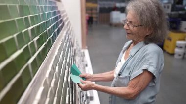 Yürüteç sahibi yaşlı bir kadın, ev dekorasyonu ve restorasyon projeleri için seçenekleri göz önüne alarak, bir hırdavatçıda bir dizi boya renk örneklerine göz atıyor..