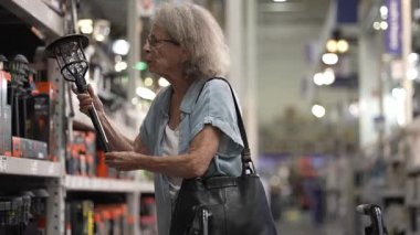 Yürüteç kullanan yaşlı bir kadın hırdavatçıdaki malzemeleri elden geçiriyor, projesi için çeşitli araçları ve ışıklandırma seçeneklerini dikkatlice inceliyor..
